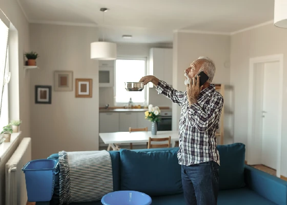 old person holding saucepan under leak calling on the phone